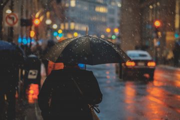 Une personne de dos marchant sous la pluie, abritée sous un parapluie dans une grande ville la nuit.