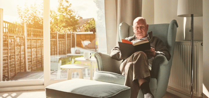 Un homme âgé, confortablement installé pour lire dans un fauteuil bleu-vert, disposé dans une chambre lumineuse avec vue sur le jardin.