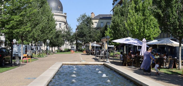 Une fontaine miroir d'eau sur une place de centre-ville arborée, avec terrasses de café et restaurants.