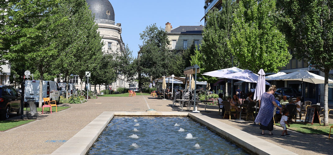 Une fontaine miroir d'eau sur une place de centre-ville arborée, avec terrasses de café et restaurants.