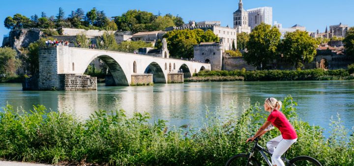 Une femme à vélo sur des berges de rivière, une ville historique touristique en arrière plan.