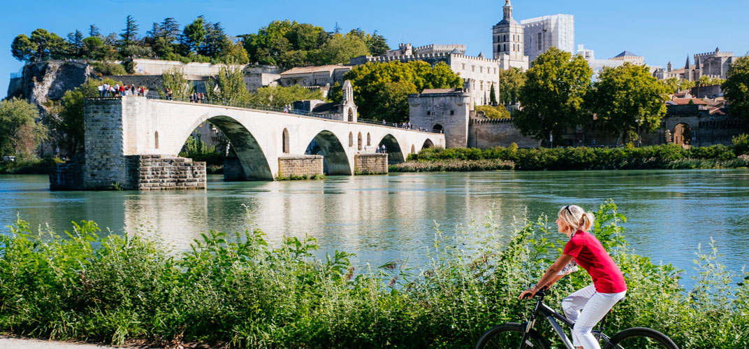 Une femme à vélo sur des berges de rivière, une ville historique touristique en arrière plan.