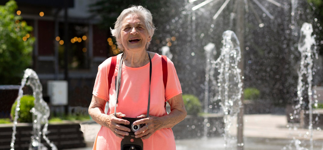 Ville intergénérationnelle : une femme âgée, souriant appareil photo en main devant une fontaine.