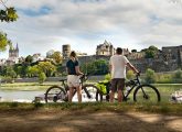 Un couple de cycliste sur les berges d'un fleuve, les fortifications d'une ville sur l'autre rive. Itw de Jérôme Barrier. ©Istock.