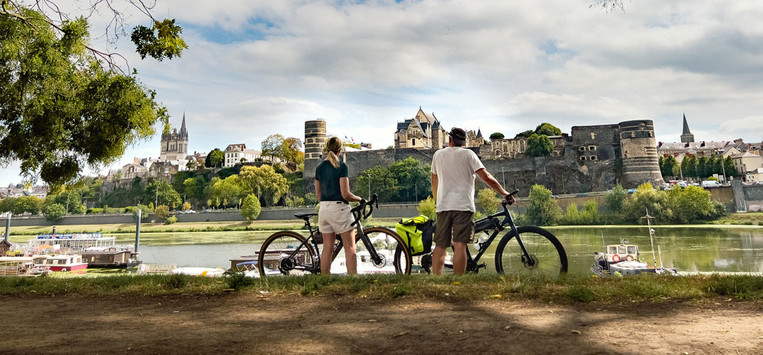 Un couple de cycliste sur les berges d'un fleuve, les fortifications d'une ville sur l'autre rive. Itw de Jérôme Barrier. ©Istock.