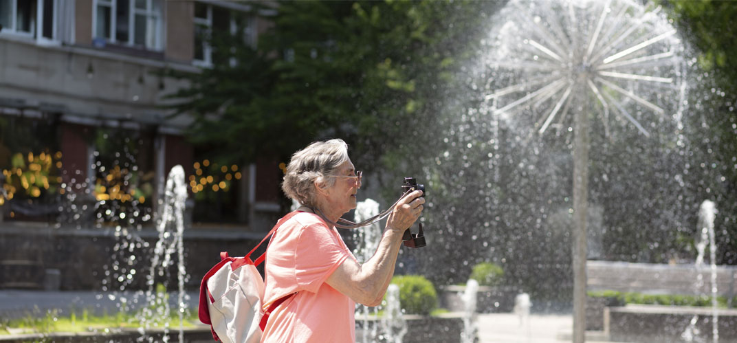 Economies d'eau en ville : Une femme âgée photographiant des fontaines et jeux d'eau sur une place par une chaude journée d'été.